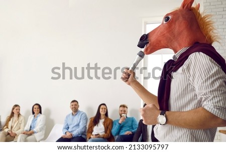 Bizarre speaker making funny absurd presentation in front of happy audience. Man wearing silly wacky horse mask holding microphone and giving lecture during business training, conference or seminar