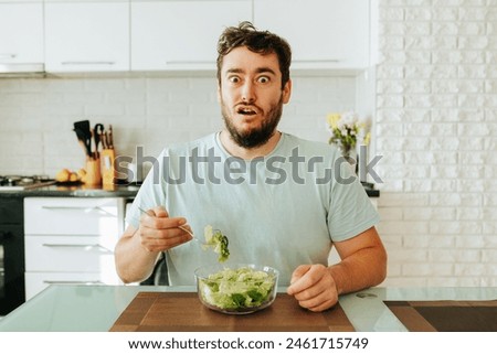 bizarre Young man expression horror on face parted mouth opened eyes wide hold a fork with salad in hand. A man should eat salad to be healthy, but he is tired of such food. Stop diet.