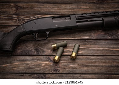 A black 12-gauge pump-action shotgun and ammo are on a wooden table. A smooth-bore weapon with a plastic stock. Dark brown background.