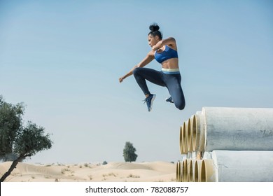 Black African American Athletic Woman Jumps Over And Leaps From Construction Pipes Wearing Sports Outfit In A Parkour Or Extreme Fitness Competition Wearing A Sports Outfit.