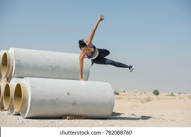 Black African American Athletic Woman Jumps Over And Leaps From Construction Pipes Wearing Sports Outfit In A Parkour Or Extreme Fitness Competition Wearing A Sports Outfit.