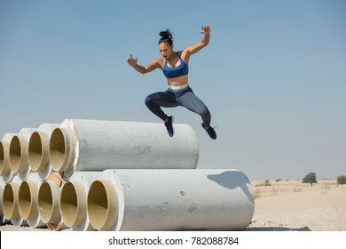 Black African American Athletic Woman Jumps Over And Leaps From Construction Pipes Wearing Sports Outfit In A Parkour Or Extreme Fitness Competition Wearing A Sports Outfit.