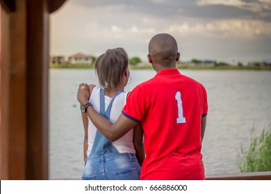 Black African American Couple Standing With Backs To Camera Looking At Lake View With Man's Arm Around Woman's Shoulder