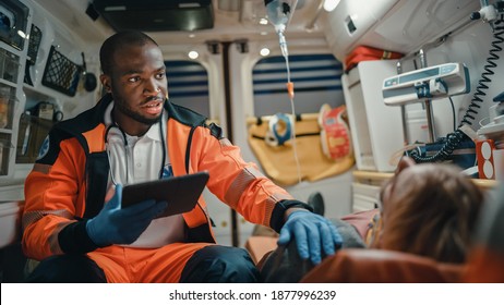 Чернокожий афроамериканский фельдшер EMS использует планшетный компьютер для заполнения анкеты для раненого пациента по дороге в больницу. Помощник скорой медицинской помощи работает в машине скорой помощи.