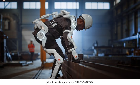 Black African American Engineer Is Testing A Futuristic Bionic Exoskeleton And Picking Up Metal Objects In A Heavy Steel Industry Factory. Contractor Is Heavy Lifting Steel Parts In A Powered Shell.