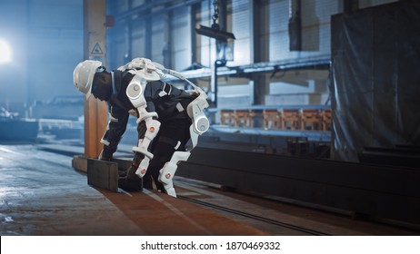 Black African American Engineer Is Testing A Futuristic Bionic Exoskeleton And Picking Up Metal Objects In A Heavy Steel Industry Factory. Contractor Is Heavy Lifting Steel Parts In A Powered Shell.