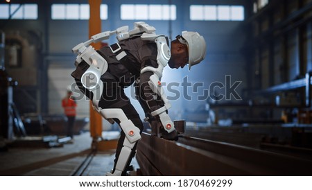 Black African American Engineer is Testing a Futuristic Bionic Exoskeleton and Picking Up Metal Objects in a Heavy Steel Industry Factory. Contractor is Heavy Lifting Steel Parts in a Powered Shell.
