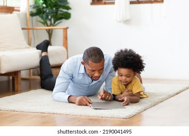 Black African American Father And Little Son Lying On Floor And Reading A Book Together At Home.