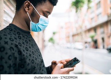 Black African American Guy On The Street With A Blue Face Mask Looking At His Cell Phone Protecting Himself From The Covid-19 Coronavirus Pandemic