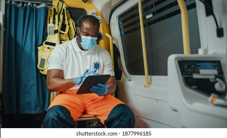 Black African American Paramedic In Face Mask Using Tablet Computer While Riding In An Ambulance Vehicle For An Emergency. Emergency Medical Technicians Outside The Healthcare Hospital.