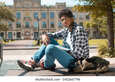 A Black African American Student In Glasses With Books Sits Near A College.