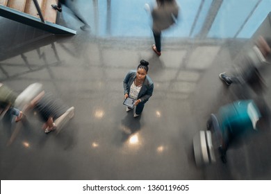 Black African Business Woman Standing In The Lobby Of An Airport Looking Up At The Camera While People Are Walking Past In A Blur