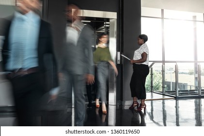 Black African Business Woman Standing With Paperwork, Waiting To Enter A Busy Elevator Lift. With People Walking Out In A Blur, Showing Motion.