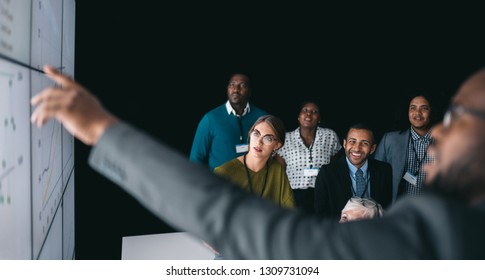 Black African Businessman Giving A Presentation At A Business Conference. Large Video Screen With Charts And Graphs Next To Him