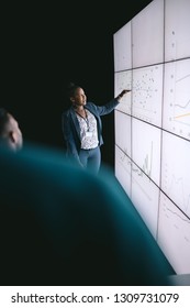 Black African Businesswoman Giving A Presentation At A Business Conference. Pointing At A Large Video Screen With Charts And Graphs Next To Her In A Dark Room