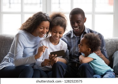 Black African Family Of Mom Dad Parents And Happy 2 Mixed Race Little Kids Sit Together On Sofa Laughing Looking At Smartphone Taking Selfie, Making Video Call, Watching Funny Video On Phone At Home