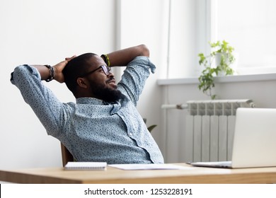 Black African Millennial Businessman Take Break During Workday Relaxing Sitting On Chair At Office Desk Hold Hands Behind Head Looking At Window Thinking Feels Good. Stress Relief Daydreaming Concept