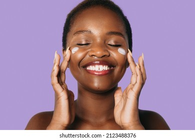 Black African Model Applying Moisturiser Face Cream To Her Cheek Isolated Over Purple Background. Toothy Smile. Facial Skincare. Happy Face.