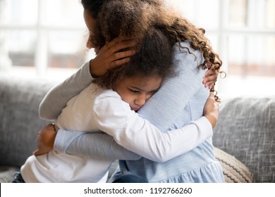 Black African mother embrace little preschool frustrated kid sitting on couch together at home. American loving mother supports disappointed daughter sympathizing, making peace after scolding concept