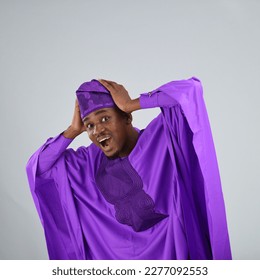 Black African Nigeria Yoruba wearing purple agbada traditional clothes holding his head being surprise - Powered by Shutterstock