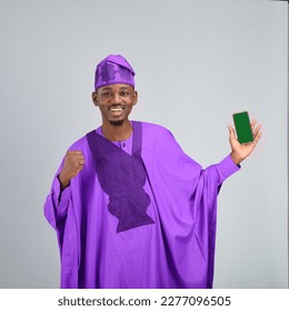 Black African Nigeria Yoruba wearing purple agbada traditional clothes holding his fist and his phone happy excited - Powered by Shutterstock