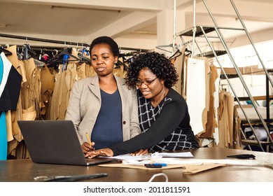 A black African Zimbabwean woman working with colleague processing paperwork with a laptop in textile factory with clothing patterns in the background wearing corporate wear