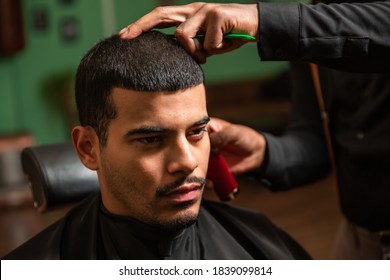 A black African-American barber cuts and shaves the hair of a Hispanic Latino man with a goatee and mustache with an electric razor in a barber shop.
