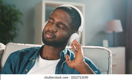 Black afro american man wearing white headphones and listening to music on his smart phone at home. Slow motion