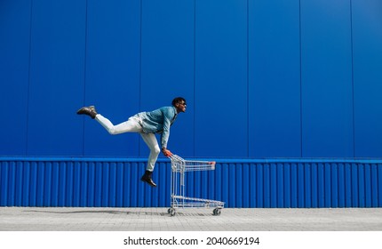 black afro guy shopping with a shopping cart, guy having fun and jumping, runs shopping near the mall, young black student on background of a blue building, black friday, sale