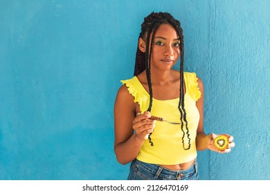 black afro latin american healthy girl with braids eating a kiwi with a spoon. Yellow shirt and blue jeans skirt. Blue wall in the background, turqouise. Beautiful skinny woman ,green yellow make up - Powered by Shutterstock