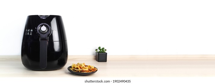 a black air fryer or oil free fryer appliance is on the wooden table in the kitchen with deep fried banan chips and small plant in the pot on white cement wall background during dinner in the party - Shutterstock ID 1902490435
