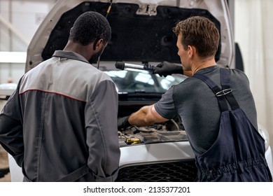 black and american or caucasian men can fix anything. handsome car mechanics in overalls uniform checking the engine under hood in modern clean car service station, workshop. rear view