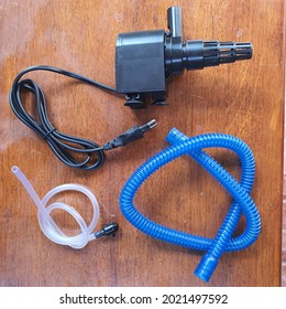 Black Aquarium Submersible Pump, Blue Hose And Transparent Tube On Wooden Background. Pump For Water Circulation And Wave Maker. Fish Aquaculture Tank And Water System.