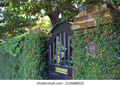 Black arched gate doorway entrance to garden enclosed by a plant-covered stone brick wall.