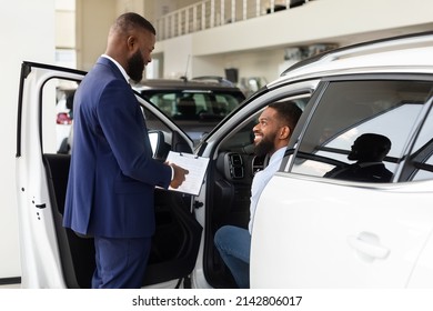 Black Auto Salon Manager Consulting Male Customer Choosing New Car, Professional Salesperson Advertising Vehicle Characteristics To Excited African American Man Sitting Inside Of Automobile