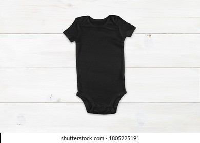 Black Baby Onesie Resting Atop A Modern White Wood Background.