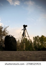 black backpack and a professional camera on a tripod on the ground or stone, their silhouettes against the bright evening sky with clouds and dark tree crowns, preparation for a photo shoot