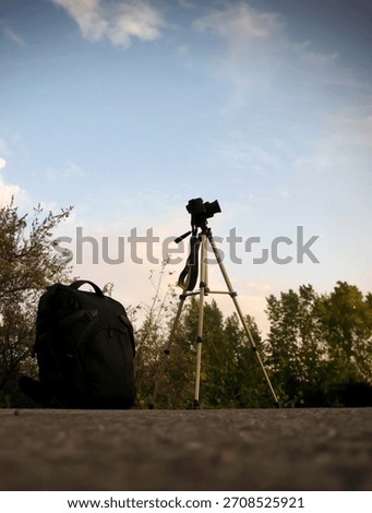 black backpack and a professional camera on a tripod on the ground or stone, their silhouettes against the bright evening sky with clouds and dark tree crowns, preparation for a photo shoot