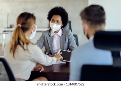 Black bank manager wearing a face mask and using touchpad while having a meeting with a couple in the office.  - Powered by Shutterstock