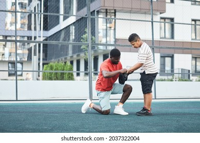 Black Baseball Player Being Down On One Knee While Putting Baseball Glove To Son Before Game At Outdoor Playground