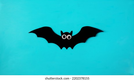 A Black Bat With White Eyes On Blue Background. Spooky Minimal Creative Concept For Halloween Wallpaper Or Web Banner Or Advertisement. Flat Lay