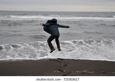 Black Beach. Black Sand. Overcast Sky. The Guy Jumps High, Arms Outstretched. Young Guy On The Beach Near The Ocean Jumping. A Young Guy On The Beach In A Warm Jacket. Silhouette