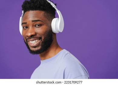 Black bearded man smiling while listening music with headphones isolated over purple background