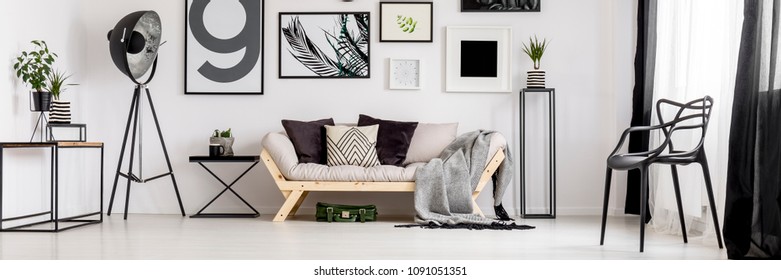 Black Big Studio Lamp Placed In The Corner Of White Living Room Interior With Wooden Couch, Many Posters And Plastic Chair