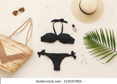 Black bikini swimsuit, straw boater hat, wicker beach bag, sunglasses, gold necklace, rings, tropical palm leave on beige background. Woman's swimwear and beach accessories. Flat lay, top view, outfit