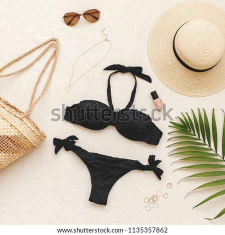 Black bikini swimsuit, straw boater hat, wicker beach bag, sunglasses, gold necklace, rings, tropical palm leave on beige background. Woman's swimwear and beach accessories. Flat lay, top view, outfit
