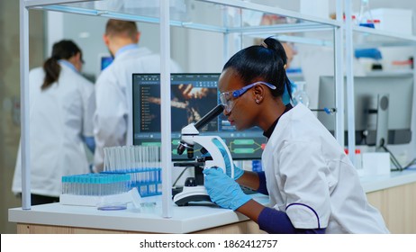 Black Biochemist Woman Sitting In Modern Equipped Lab Analysing Blood Tests. Multiethnic Team Examining Virus Evolution Using High Tech For Scientific Research Of Treatment Development Against Covid19