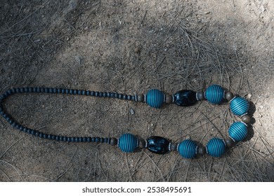 black and blue beaded necklace with round and textured shape, lies on sandy ground with dry twigs. - Powered by Shutterstock