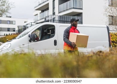 Black blue-collar worker - package delivery person - in front of white truck with cardboard package. Clothing - red jacket with gray sleeves and black hat. Blurred tall grass in the foreground. High - Powered by Shutterstock