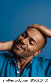 Black Bold Man Wearing Shirt Smiling While Holding His Head Isolated Over Blue Background